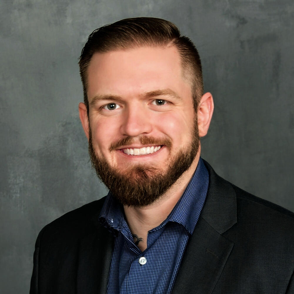 Professional headshot of a man with short dark hair and a beard, wearing a navy shirt and dark blazer, smiling against a gray studio background.